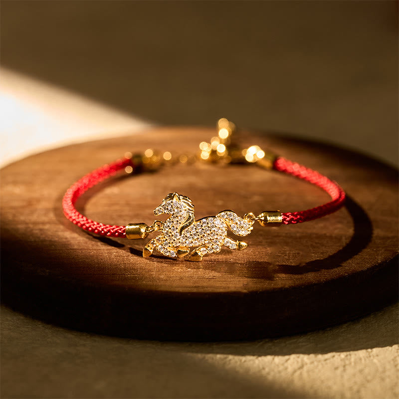 Gold bracelet with a horse charm on a red string, placed on a wooden surface.