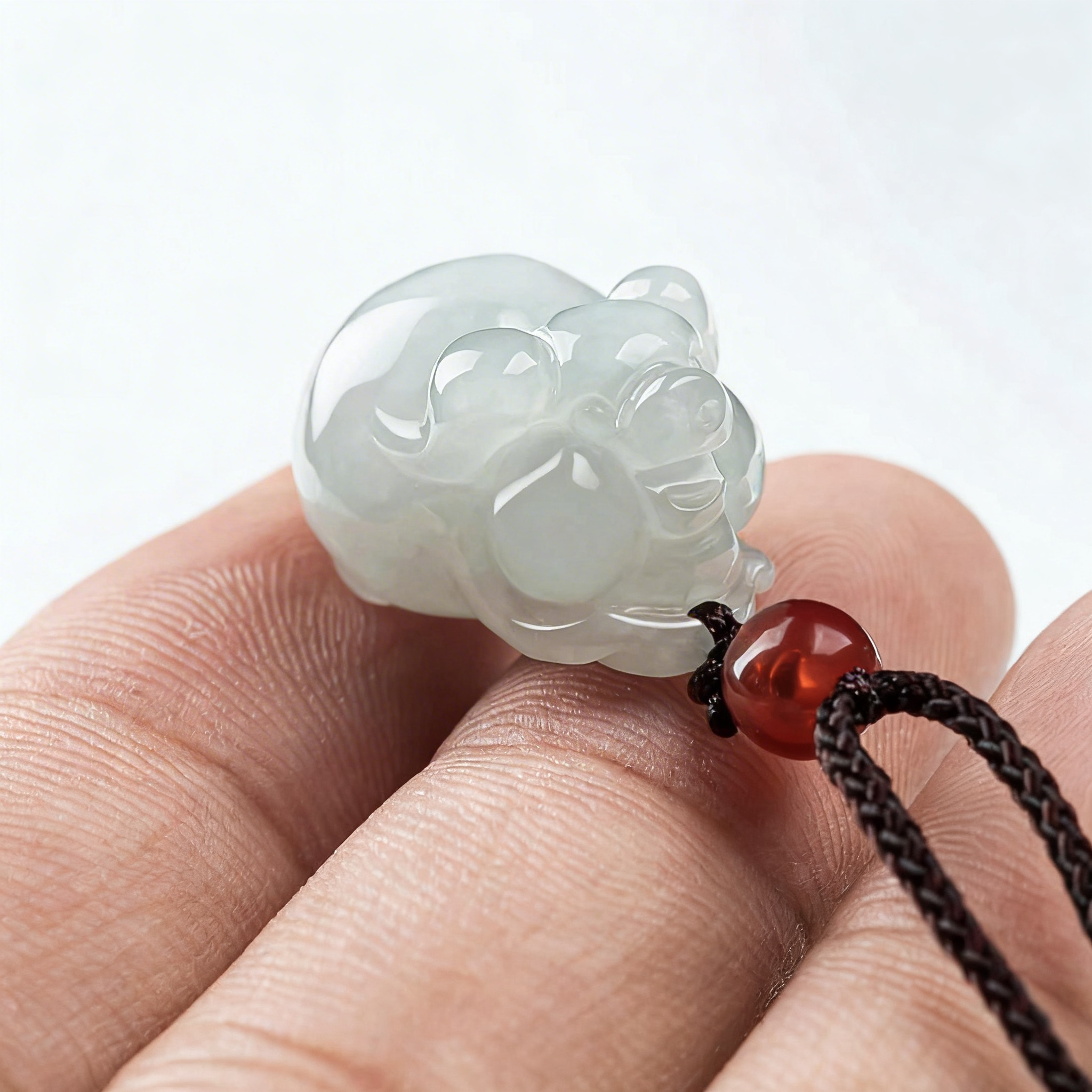 White jade pendant shaped like a bear held between fingers with a red bead on a black cord.