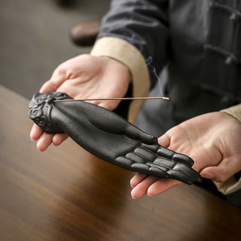 Person holding a black incense holder shaped like a hand with an incense stick.