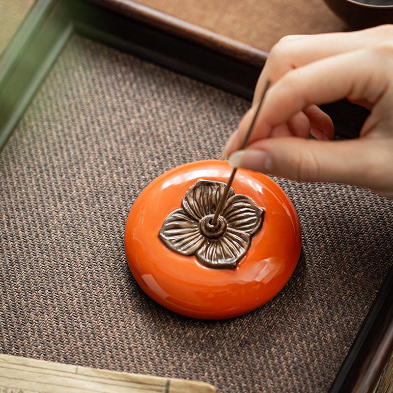Hand holding a small tool near an orange ceramic object with a floral design on a textured surface.