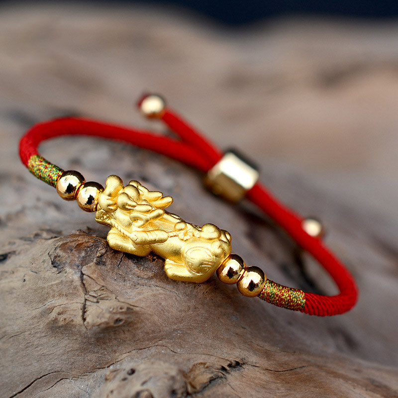 Gold bracelet with a dragon charm on a red cord, placed on a wooden surface.