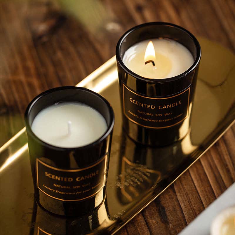 Two scented candles in black containers on a reflective surface with a blurred background.