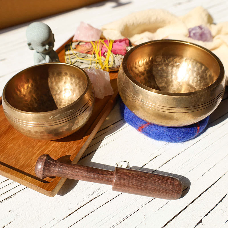 Two brass singing bowls with a wooden mallet on a wooden surface.