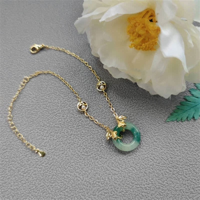 Gold bracelet with a green jade charm on a gray surface with white flowers.