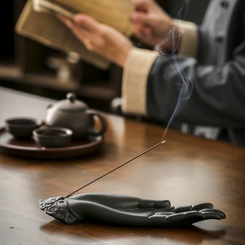 Incense stick in a hand-shaped holder with a teapot and cups on a wooden table